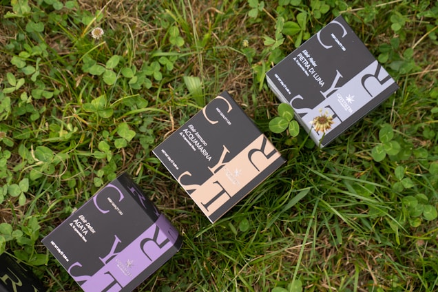 Several boxes with elegant designs labeled with different product names are placed on a patch of green grass and clover. The boxes have distinct typography and color schemes, including shades of purple, blue, and beige.
