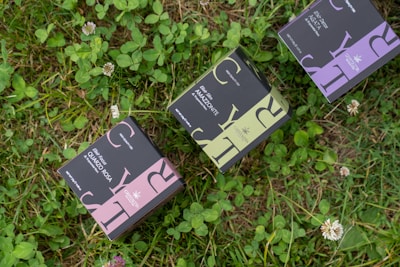 Three small rectangular boxes with elegant packaging design featuring large letters and intricate patterns are placed on lush green grass with scattered clover and small white flowers.