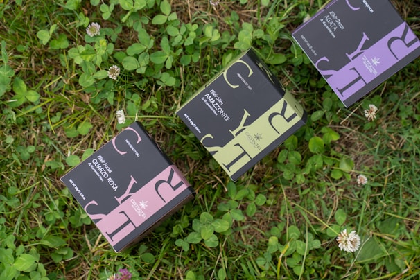 Three small rectangular boxes with elegant packaging design featuring large letters and intricate patterns are placed on lush green grass with scattered clover and small white flowers.