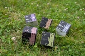 Several product boxes are placed on a grassy field. The boxes are designed with different color schemes and feature bold typography. They appear to contain cosmetics or personal care items.