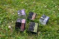 Several product boxes are placed on a grassy field. The boxes are designed with different color schemes and feature bold typography. They appear to contain cosmetics or personal care items.