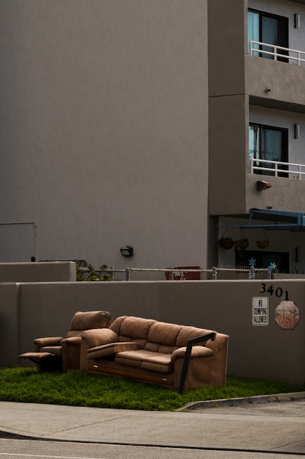 A brown sofa set, including a couch and an armchair, is placed on a patch of grass next to a sidewalk. The setting is in front of a gray concrete building with apartment balconies. A 'No Dumping Allowed' sign is visible on the wall behind the furniture.