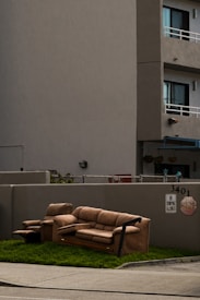 A brown sofa set, including a couch and an armchair, is placed on a patch of grass next to a sidewalk. The setting is in front of a gray concrete building with apartment balconies. A 'No Dumping Allowed' sign is visible on the wall behind the furniture.