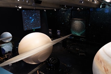 A museum exhibit featuring large models of planets, including Saturn with its rings, and other celestial objects displayed against a backdrop of stars. Screens present images of a galaxy and a star cluster.