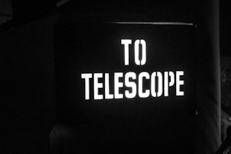 A black-and-white illuminated sign with the words 'TO TELESCOPE' displayed in large, bold letters. The sign is likely pointing towards an observatory or a location where a telescope is set up. The surroundings are dark, suggesting a night setting or a dimly lit area.