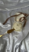 A small, white frosted cake with a single lit candle on top sits in an open white box adorned with a brown ribbon. The cake rests on a silky, silver fabric that creates a luxurious background. A gold cake server lies next to the cake, adding a touch of elegance.