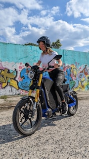 A person wearing a helmet and sunglasses rides a blue and black electric motorcycle with yellow front forks. The background features a colorful graffiti-covered wall under a blue sky with scattered clouds.
