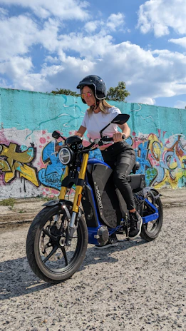 Smiling rider wearing gloves and helmet, standing next to an electric motorcycle under soft blue lighting.