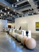 A modern gallery space with high ceilings and exposed lighting fixtures. There are several people browsing and taking photos of the art installations. The main installation features large spherical stones and mirrored panels arranged in a line on the polished concrete floor. The walls are adorned with various artworks, including abstract pieces. The overall ambiance is clean and contemporary.