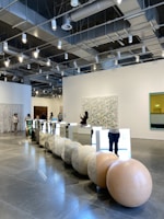 A modern gallery space with high ceilings and exposed lighting fixtures. There are several people browsing and taking photos of the art installations. The main installation features large spherical stones and mirrored panels arranged in a line on the polished concrete floor. The walls are adorned with various artworks, including abstract pieces. The overall ambiance is clean and contemporary.