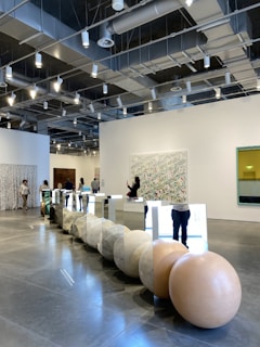 A modern gallery space with high ceilings and exposed lighting fixtures. There are several people browsing and taking photos of the art installations. The main installation features large spherical stones and mirrored panels arranged in a line on the polished concrete floor. The walls are adorned with various artworks, including abstract pieces. The overall ambiance is clean and contemporary.