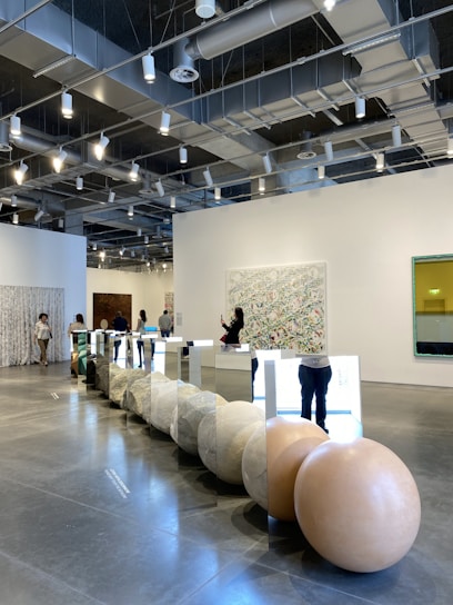 A modern gallery space with high ceilings and exposed lighting fixtures. There are several people browsing and taking photos of the art installations. The main installation features large spherical stones and mirrored panels arranged in a line on the polished concrete floor. The walls are adorned with various artworks, including abstract pieces. The overall ambiance is clean and contemporary.