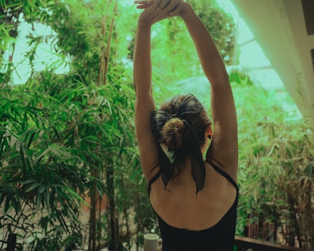 A person with a black bow in their hair is stretching their arms overhead while surrounded by lush green foliage. The setting suggests a vibrant, natural environment, possibly indoors.