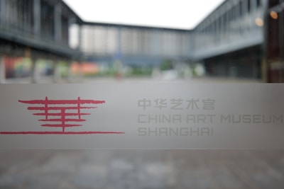 A glass window with a frosted design displays text and a logo in red, with the words 'China Art Museum Shanghai' visible. The background shows an outdoor view of modern architectural structures, slightly blurred.