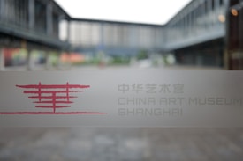 A glass window with a frosted design displays text and a logo in red, with the words 'China Art Museum Shanghai' visible. The background shows an outdoor view of modern architectural structures, slightly blurred.