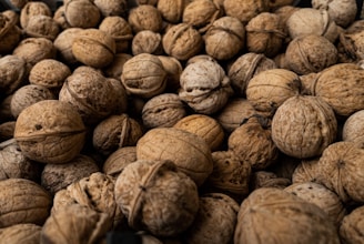 Numerous walnuts with their shells intact are scattered closely together, creating a textured surface. The image captures the natural, rustic appearance of the nuts in varying shades of brown.