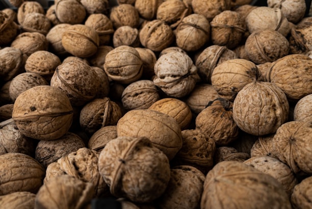 Numerous walnuts with their shells intact are scattered closely together, creating a textured surface. The image captures the natural, rustic appearance of the nuts in varying shades of brown.