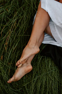 A serene woman sitting barefoot on soft grass in a teal and sand-colored natural setting, eyes closed in peaceful reflection.