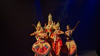 Five dancers in traditional attire perform a classical Indian dance on a dimly lit stage. The performers are adorned with intricate headpieces and jewelry, showcasing vibrant costumes in shades of red, pink, and gold, with elaborate patterns. The dancers are poised in an elegant formation, reflecting precision and grace.