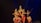 Five dancers in traditional attire perform a classical Indian dance on a dimly lit stage. The performers are adorned with intricate headpieces and jewelry, showcasing vibrant costumes in shades of red, pink, and gold, with elaborate patterns. The dancers are poised in an elegant formation, reflecting precision and grace.