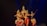 Five dancers in traditional attire perform a classical Indian dance on a dimly lit stage. The performers are adorned with intricate headpieces and jewelry, showcasing vibrant costumes in shades of red, pink, and gold, with elaborate patterns. The dancers are poised in an elegant formation, reflecting precision and grace.
