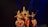 Five dancers in traditional attire perform a classical Indian dance on a dimly lit stage. The performers are adorned with intricate headpieces and jewelry, showcasing vibrant costumes in shades of red, pink, and gold, with elaborate patterns. The dancers are poised in an elegant formation, reflecting precision and grace.