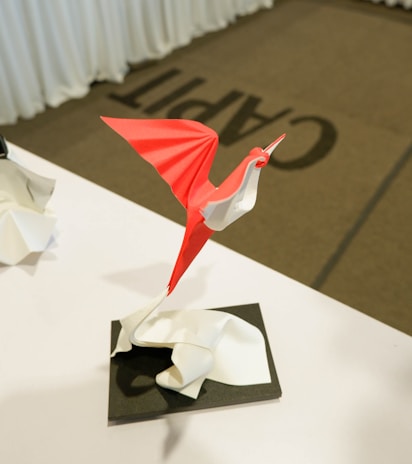 An origami sculpture resembling a bird is elegantly perched on a surface. The bird is crafted from red and white paper, with the red forming the wings and body, while the white makes up the neck and head. It appears to be in a poised, dynamic position, suggesting motion. The background includes a carpet with large, partially visible lettering.