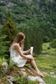 A woman with long hair sits on a moss-covered rock in a lush, green forest holding a tube of product. She is wearing a sleeveless white dress and appears to be in a serene, natural setting surrounded by trees and greenery.