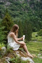 A woman with long hair sits on a moss-covered rock in a lush, green forest holding a tube of product. She is wearing a sleeveless white dress and appears to be in a serene, natural setting surrounded by trees and greenery.