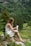 A woman with long hair sits on a moss-covered rock in a lush, green forest holding a tube of product. She is wearing a sleeveless white dress and appears to be in a serene, natural setting surrounded by trees and greenery.