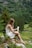 A woman with long hair sits on a moss-covered rock in a lush, green forest holding a tube of product. She is wearing a sleeveless white dress and appears to be in a serene, natural setting surrounded by trees and greenery.