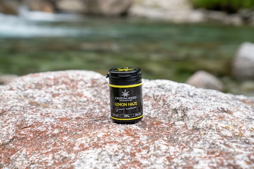 A container labeled 'Lemon Haze' from Crystalweed sits on a large, textured rock surface. The background features a flowing, blurred body of water with a greenish hue, and there are additional rocks partially visible.