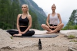 Two people are meditating outdoors in a serene natural setting, sitting cross-legged on yoga mats on a rocky surface. In the foreground, there is a small bottle with a dropper, possibly containing essential oil or a natural remedy. The background features a lush, green forest and distant mountains, evoking a peaceful and tranquil atmosphere.