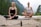 Two people are meditating outdoors in a serene natural setting, sitting cross-legged on yoga mats on a rocky surface. In the foreground, there is a small bottle with a dropper, possibly containing essential oil or a natural remedy. The background features a lush, green forest and distant mountains, evoking a peaceful and tranquil atmosphere.