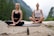 Two people are meditating outdoors in a serene natural setting, sitting cross-legged on yoga mats on a rocky surface. In the foreground, there is a small bottle with a dropper, possibly containing essential oil or a natural remedy. The background features a lush, green forest and distant mountains, evoking a peaceful and tranquil atmosphere.