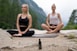 Two people are meditating outdoors in a serene natural setting, sitting cross-legged on yoga mats on a rocky surface. In the foreground, there is a small bottle with a dropper, possibly containing essential oil or a natural remedy. The background features a lush, green forest and distant mountains, evoking a peaceful and tranquil atmosphere.