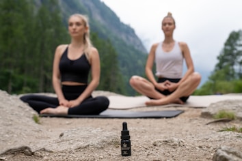 Two people are meditating outdoors in a serene natural setting, sitting cross-legged on yoga mats on a rocky surface. In the foreground, there is a small bottle with a dropper, possibly containing essential oil or a natural remedy. The background features a lush, green forest and distant mountains, evoking a peaceful and tranquil atmosphere.