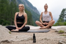 Two people are meditating outdoors in a serene natural setting, sitting cross-legged on yoga mats on a rocky surface. In the foreground, there is a small bottle with a dropper, possibly containing essential oil or a natural remedy. The background features a lush, green forest and distant mountains, evoking a peaceful and tranquil atmosphere.