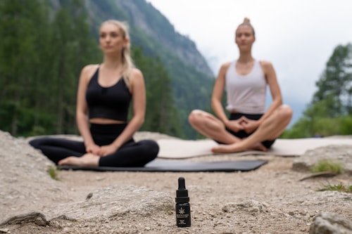 Two people are meditating outdoors in a serene natural setting, sitting cross-legged on yoga mats on a rocky surface. In the foreground, there is a small bottle with a dropper, possibly containing essential oil or a natural remedy. The background features a lush, green forest and distant mountains, evoking a peaceful and tranquil atmosphere.