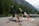 Two women are practicing yoga on mats placed on the ground beside a body of water in a mountainous forest area. They are each performing a one-legged downward dog pose, stretching their left legs upward. The surrounding landscape features a serene lake, dense green trees, and rugged mountains with mist.