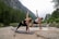 Two people are practicing yoga on mats near a serene river surrounded by lush green trees and a rocky landscape. They are both focused and stretching, with one in a black outfit and the other in a white top and black shorts.