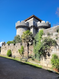 A medieval stone castle with large cylindrical towers and crenellated battlements. The structure is surrounded by lush greenery, including palm trees and bushes, and a stone wall. Benches are placed along a pathway that runs parallel to the wall under a clear blue sky, enhancing the historical and serene ambiance.