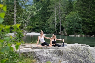 Two women are performing a yoga stretch on a rock near a serene body of water, surrounded by lush greenery and tall trees. The atmosphere is peaceful and natural.