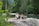 Two women are performing a yoga stretch on a rock near a serene body of water, surrounded by lush greenery and tall trees. The atmosphere is peaceful and natural.
