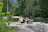 Two women are performing a yoga stretch on a rock near a serene body of water, surrounded by lush greenery and tall trees. The atmosphere is peaceful and natural.