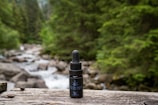 A bottle of CBD oil with a dropper sits on a wooden surface. Behind it, a blurred background reveals a forested area with lush green trees and a flowing stream over rocks.