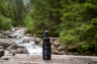 A bottle of CBD oil with a dropper sits on a wooden surface. Behind it, a blurred background reveals a forested area with lush green trees and a flowing stream over rocks.