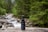 A bottle of CBD oil with a dropper sits on a wooden surface. Behind it, a blurred background reveals a forested area with lush green trees and a flowing stream over rocks.