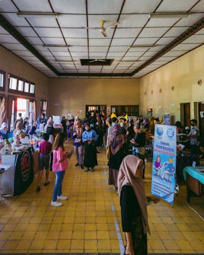 A large indoor hall with a high ceiling hosts a bustling event. Numerous people, including adults and children, are gathered, interacting with each other or attending various booths and tables set up along the walls. Informational banners are displayed, and some individuals are dressed casually while others are in more traditional attire. The atmosphere appears lively and engaging.