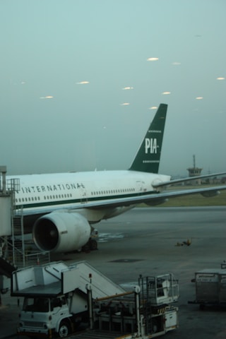 A large passenger airplane is parked at an airport terminal. The plane has the marking 'PIA' on its tail and 'INTERNATIONAL' along its fuselage. Ground support equipment, such as a mobile stairway and service vehicles, are positioned near the aircraft. The sky is overcast, giving a muted and calm atmosphere to the scene.